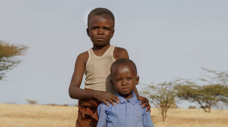 A young boy wearing yellow carries a small child in blue in the desert.