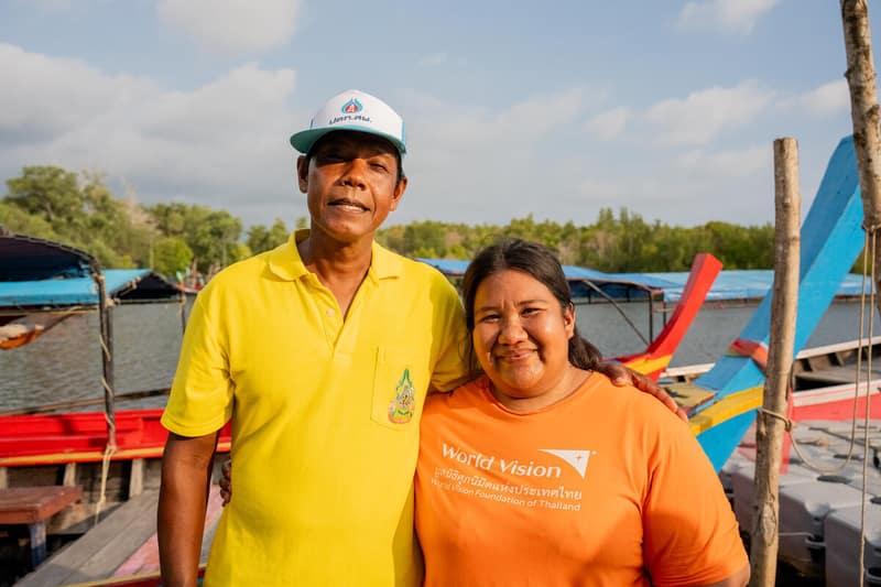 A Thai community leader and World Vision disaster committee head stand proudly in front of a restored mangrove forest lining a river.