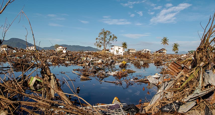 A flooded coastal landscape after the 2004 Boxing Day Tsunami, littered with branches, rubbish, damaged houses and street signs.