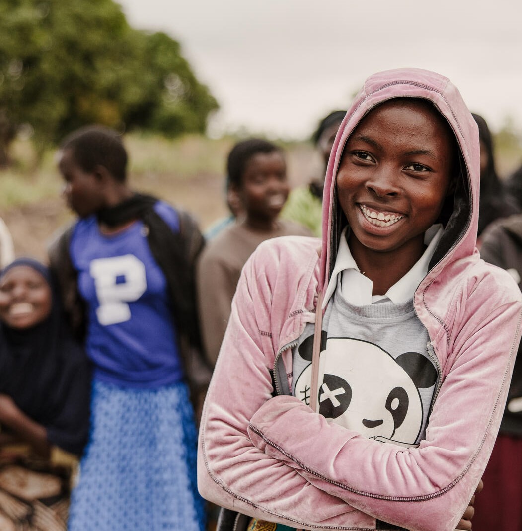 A 15-year-old girl smiles with arms crossed in a field in Malawi, surrounded by friends.