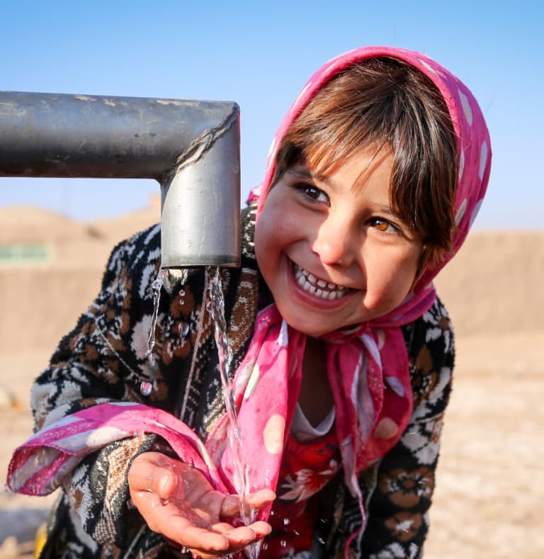 A smiling young girl wearing a head scarf, with her hands under a new tap of running water