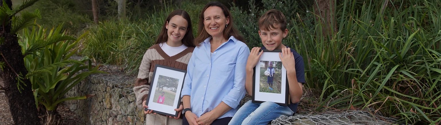 A woman and her two children standing together outdoors, proudly showing framed photos of their two sponsored children.
