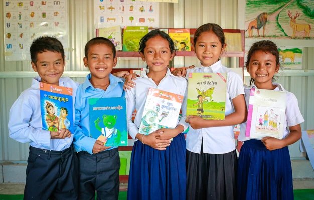 Sponsored child Nita smiling and showing a textbook with other children at a primary school in Cambodia.