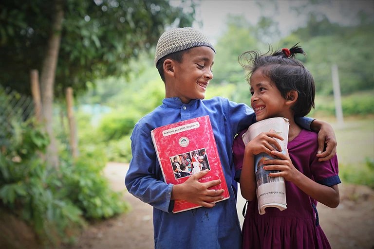 A happy boy and girl standing outdoors, holding books and smiling at each other.