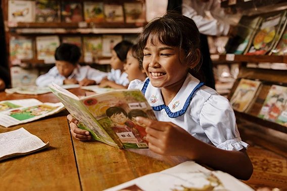 Close-up of a smiling female student in a white uniform, reading a storybook in a classroom.