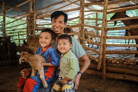 A man smiling with his two young sons and a kid goat in a farm setting.